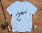 Light blue t-shirt with 'Addicted' graphic on a wooden surface with fishing equipment.

