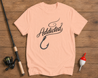 Pink t-shirt with 'Addicted' and fishing hook design on a wooden surface with fishing equipment.

