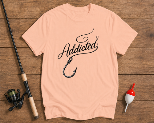 Pink t-shirt with 'Addicted' and fishing hook design on a wooden surface with fishing equipment.

