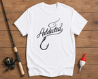 White t-shirt with 'Addicted' and fish hook design on a wooden surface with fishing equipment.

