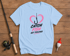 Light blue t-shirt with fishing-themed graphic on a wooden surface with fishing equipment.