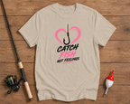 Beige t-shirt with fishing graphic and text on a wooden surface with fishing equipment.

