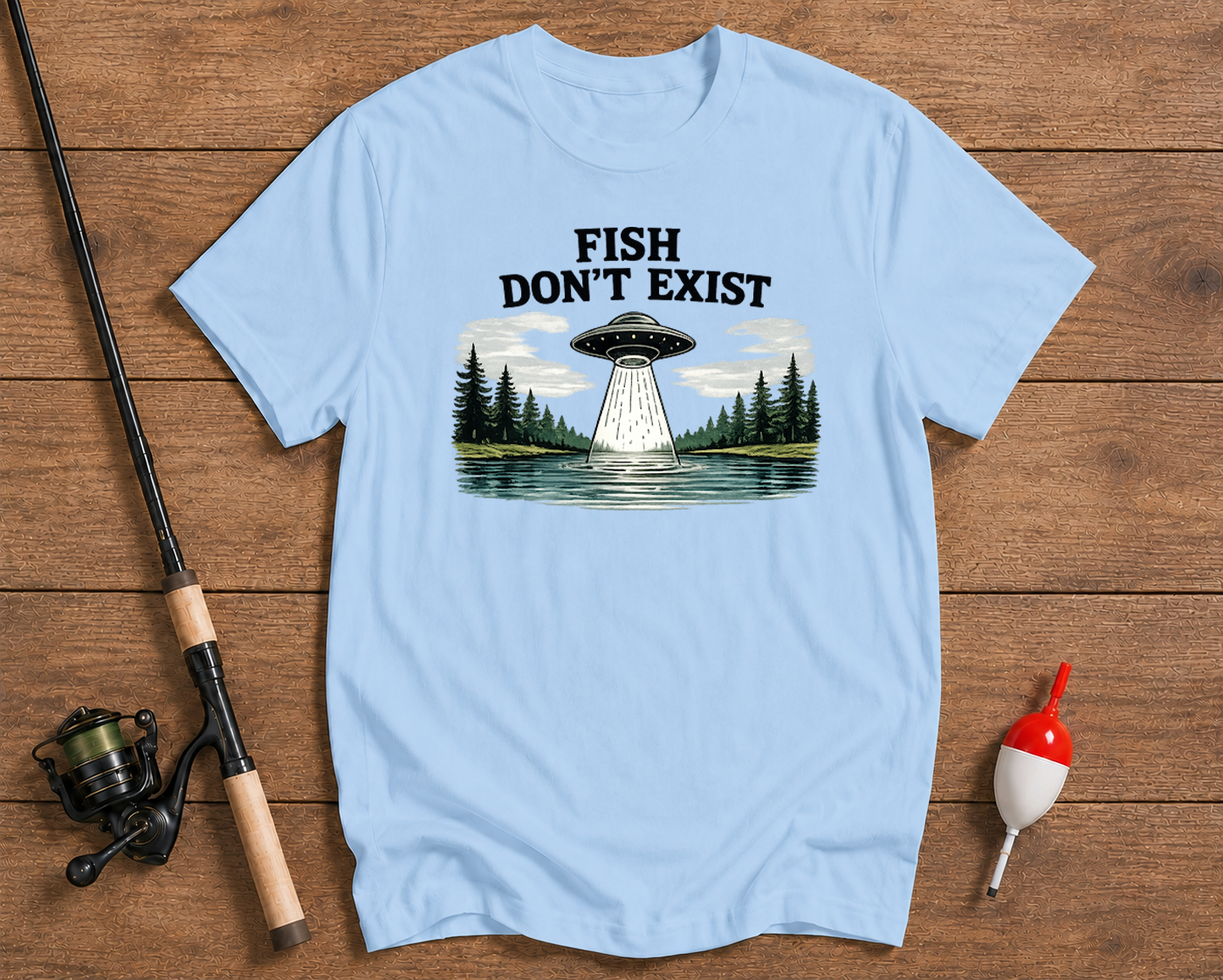 Light blue t-shirt with UFO and forest graphic, fishing rod and float on wooden surface

