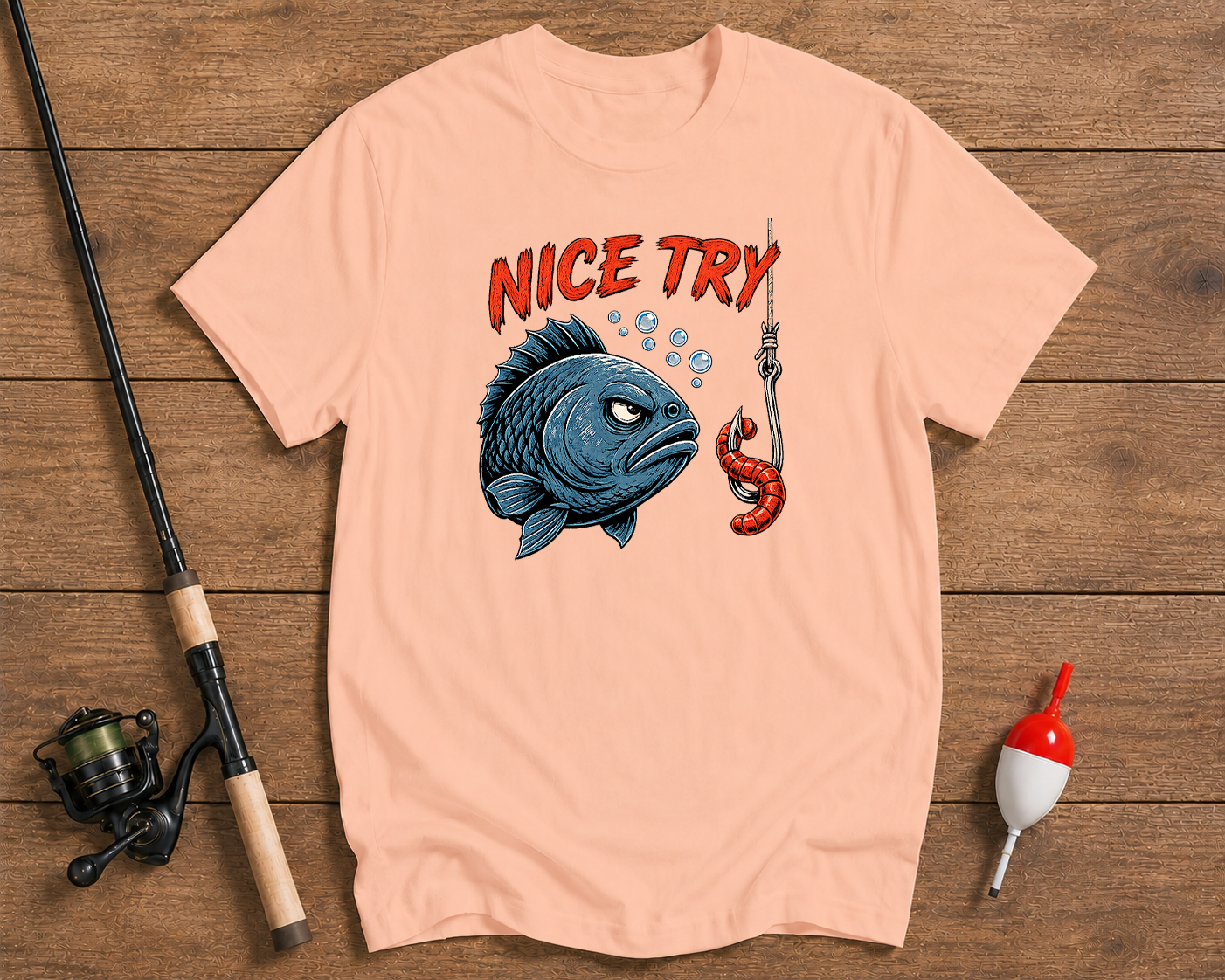 Pink t-shirt with a graphic of a fish and lobster on a wooden surface with fishing equipment.


