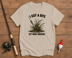 Beige t-shirt with fishing graphic and text on a wooden surface with fishing equipment.

