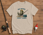 Beige t-shirt with skeleton fishing graphic and 'No Bites Just Vibes' text on a wooden surface with fishing equipment.

