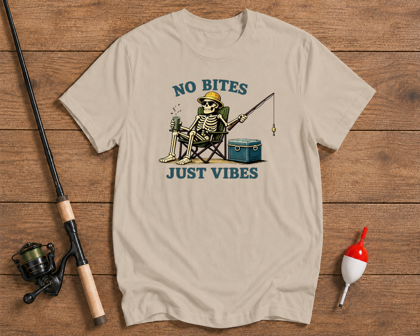 Beige t-shirt with skeleton fishing graphic and 'No Bites Just Vibes' text on a wooden surface with fishing equipment.

