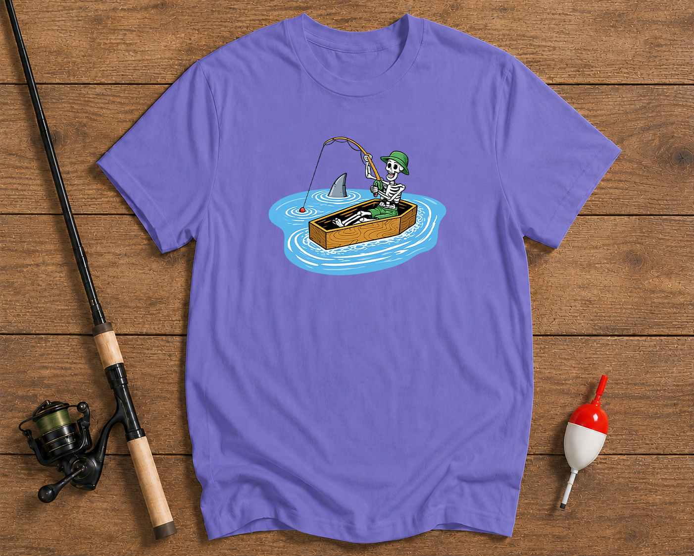 Purple t-shirt with a graphic of a skeleton fishing, placed on a wooden surface with fishing equipment.

