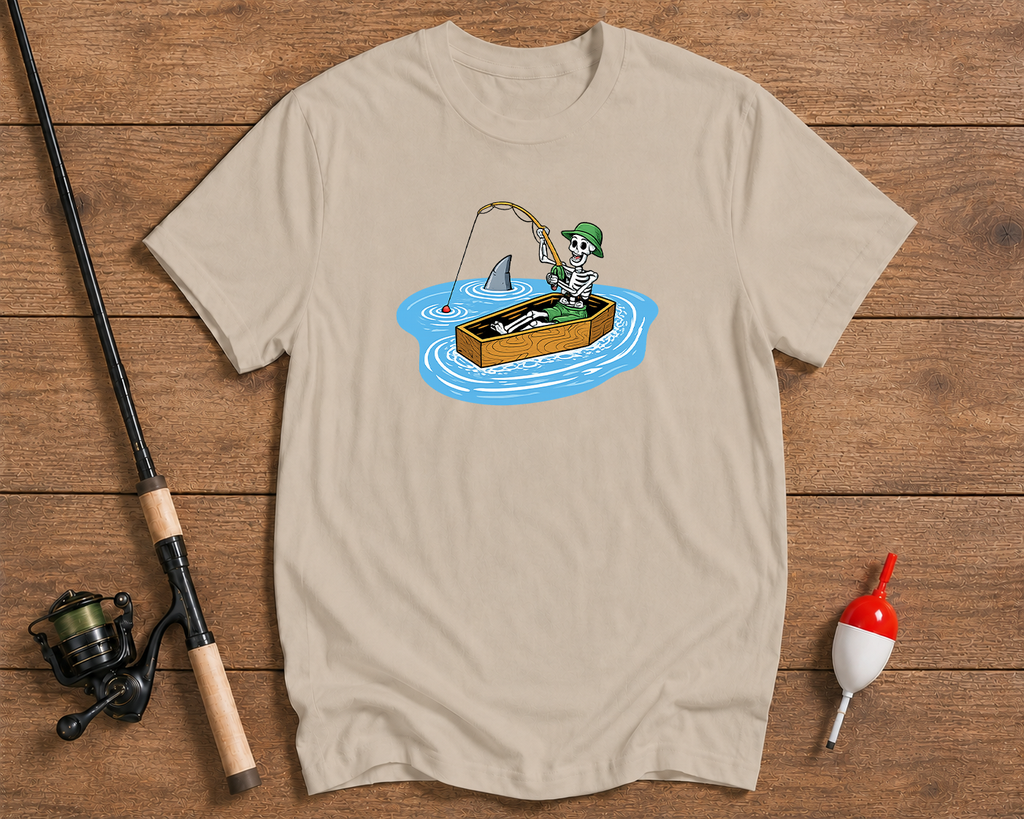 Beige t-shirt with a cartoon design of a skeleton fishing, placed on a wooden surface with fishing equipment.

