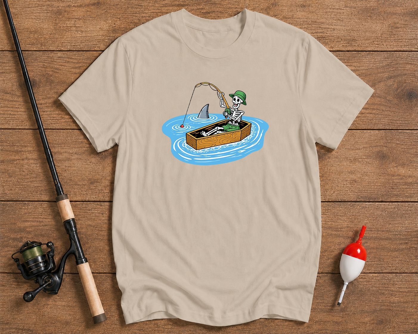 Beige t-shirt with a cartoon design of a skeleton fishing, placed on a wooden surface with fishing equipment.

