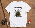 White t-shirt with fishing graphic and text on a wooden surface with fishing equipment.

