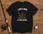 Black t-shirt with fishing graphic and text 'I got a bite (it was grass)' on a wooden surface with fishing equipment.

