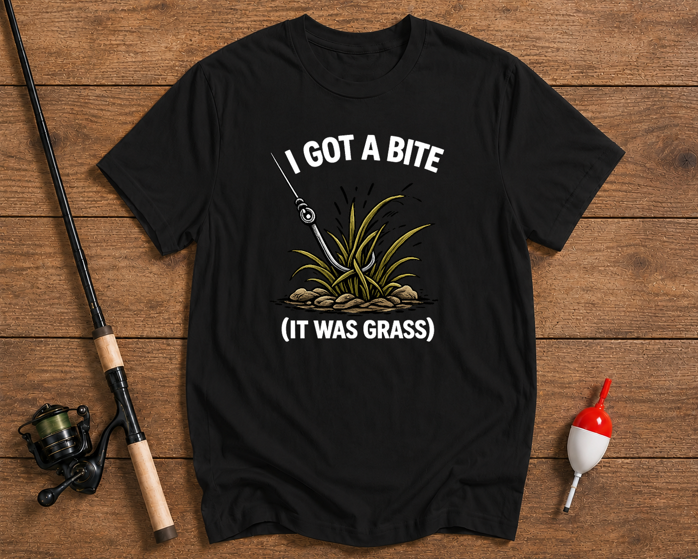 Black t-shirt with fishing graphic and text 'I got a bite (it was grass)' on a wooden surface with fishing equipment.

