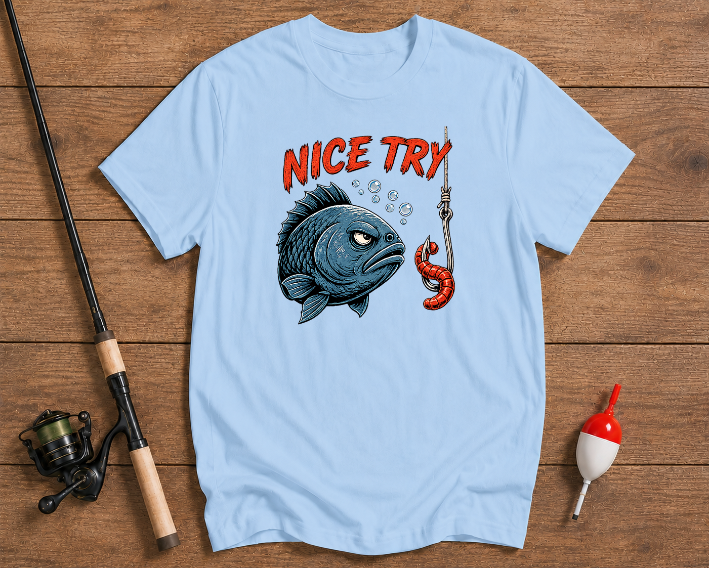 Light blue t-shirt with fishing-themed graphic and text on a wooden surface with fishing equipment.

