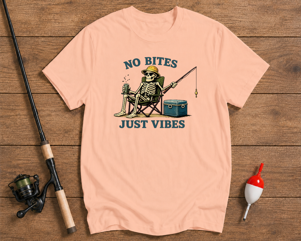 Pink t-shirt with skeleton fishing graphic and 'No Bites Just Vibes' text on a wooden surface with fishing equipment.

