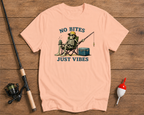 Pink t-shirt with skeleton fishing graphic and 'No Bites Just Vibes' text on a wooden surface with fishing equipment.

