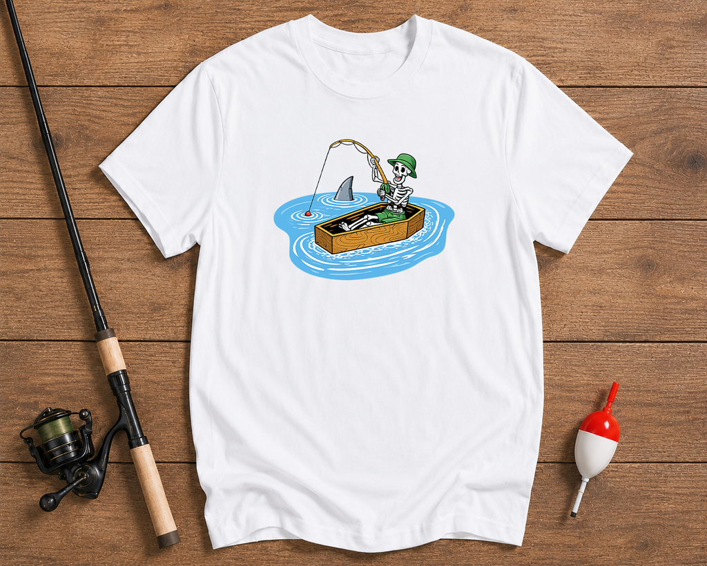 White t-shirt with a cartoon design of a person fishing from a box on a wooden surface with fishing equipment.

