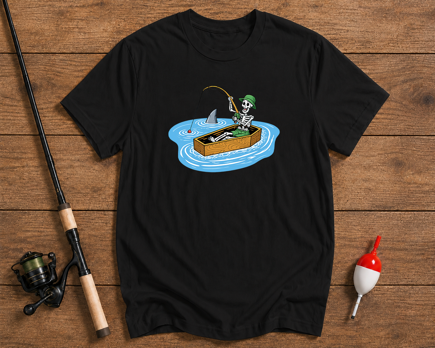 Black t-shirt with a skeleton fishing design on a wooden surface with fishing equipment.


