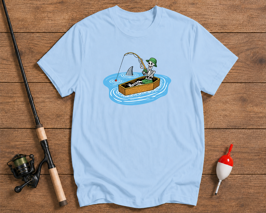 Light blue t-shirt with a graphic of a person fishing from a box with a shark, on a wooden surface with fishing equipment.

