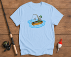 Light blue t-shirt with a graphic of a person fishing from a box with a shark, on a wooden surface with fishing equipment.

