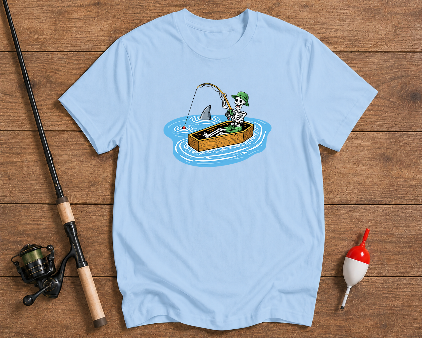 Light blue t-shirt with a graphic of a person fishing from a box with a shark, on a wooden surface with fishing equipment.

