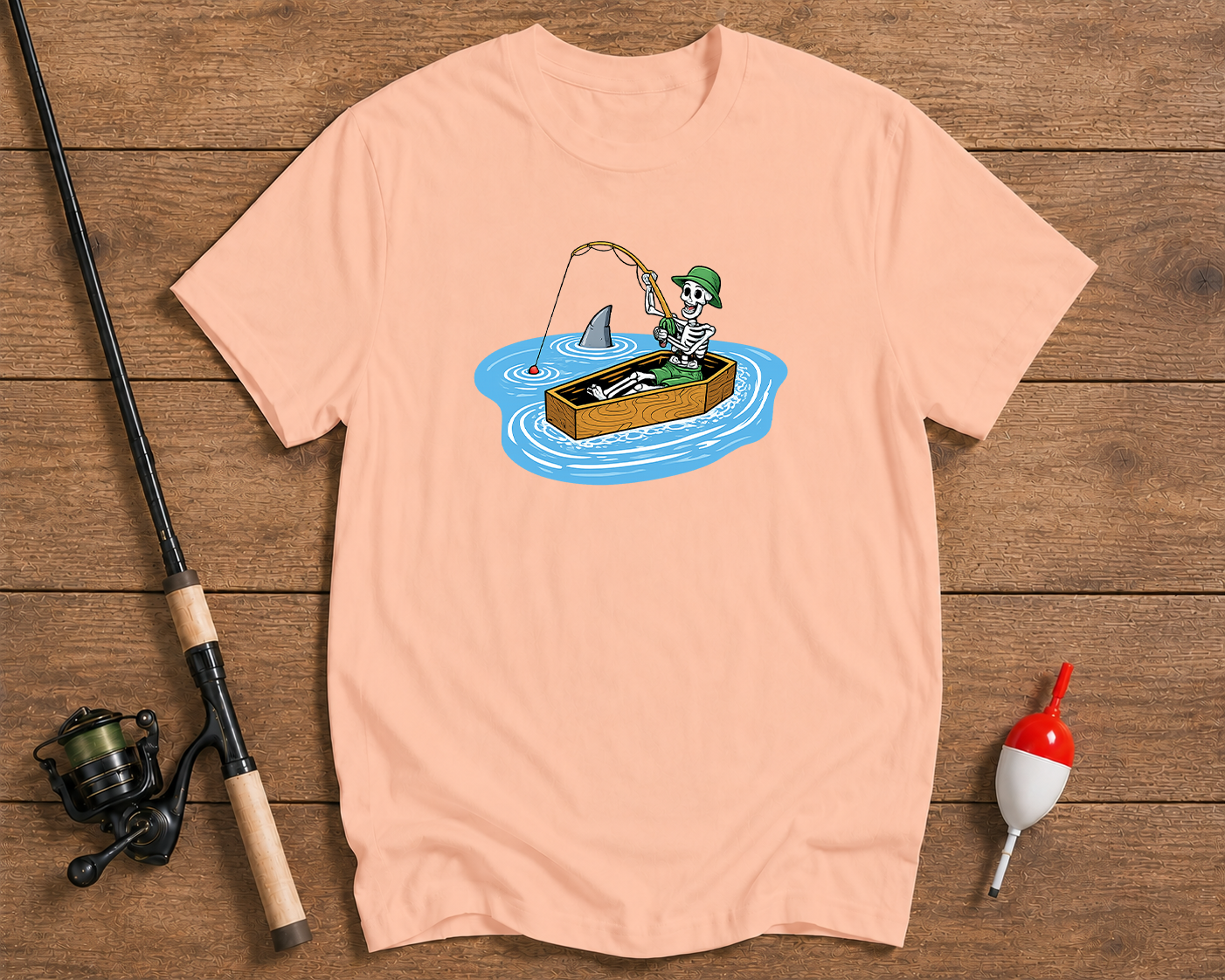Pink t-shirt with a graphic of a skeleton fishing, placed on a wooden surface with fishing equipment.

