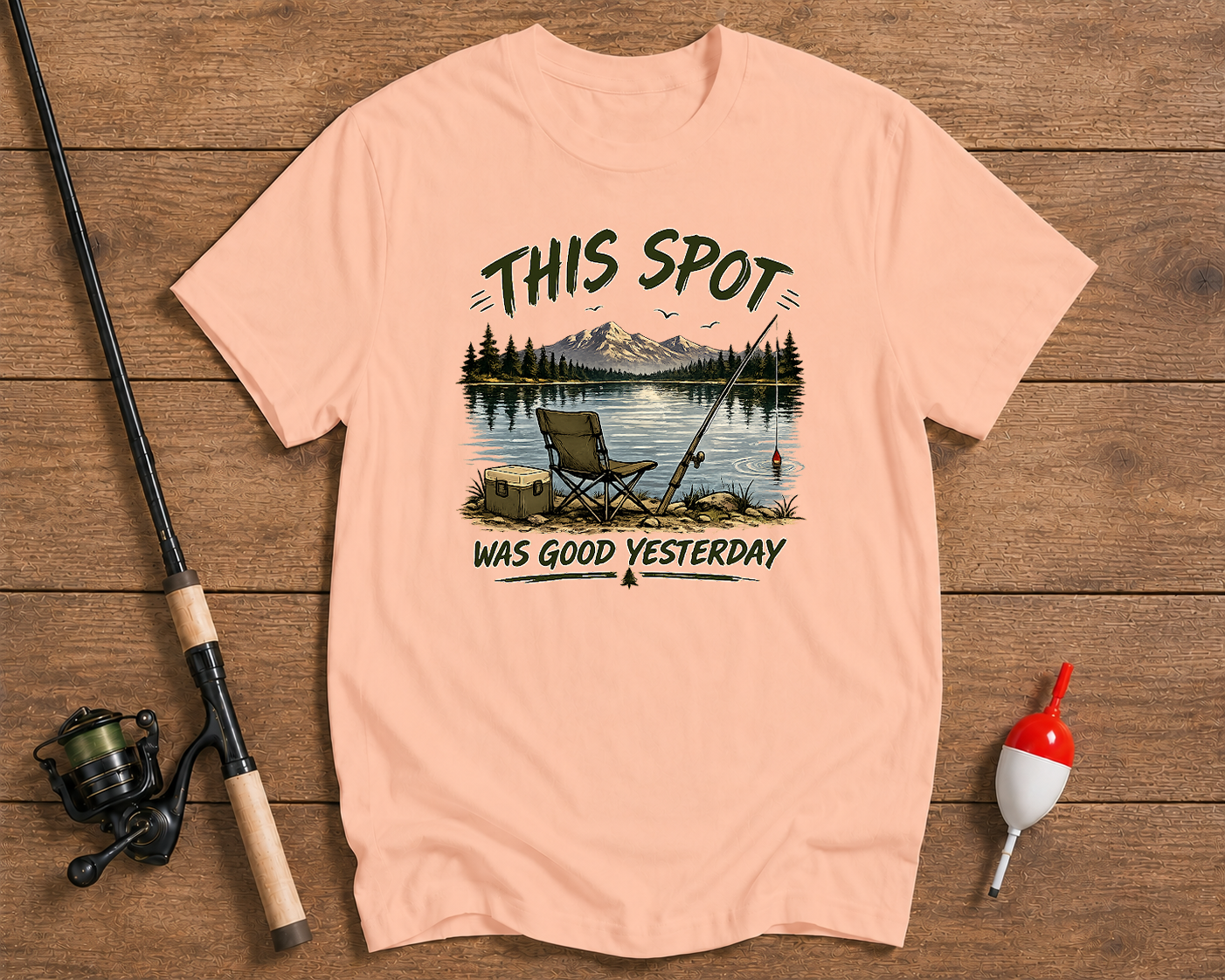 Pink t-shirt with fishing graphic and text on a wooden surface with fishing equipment.


