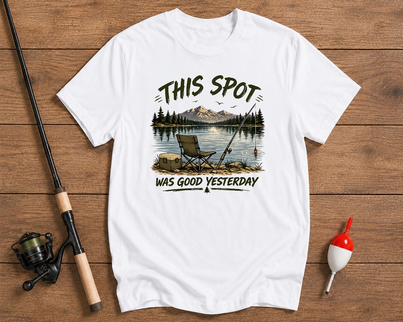 White t-shirt with fishing graphic and text on a wooden surface with fishing equipment.

