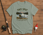 Sage green t-shirt with a fishing graphic and text on a wooden surface with fishing equipment.

