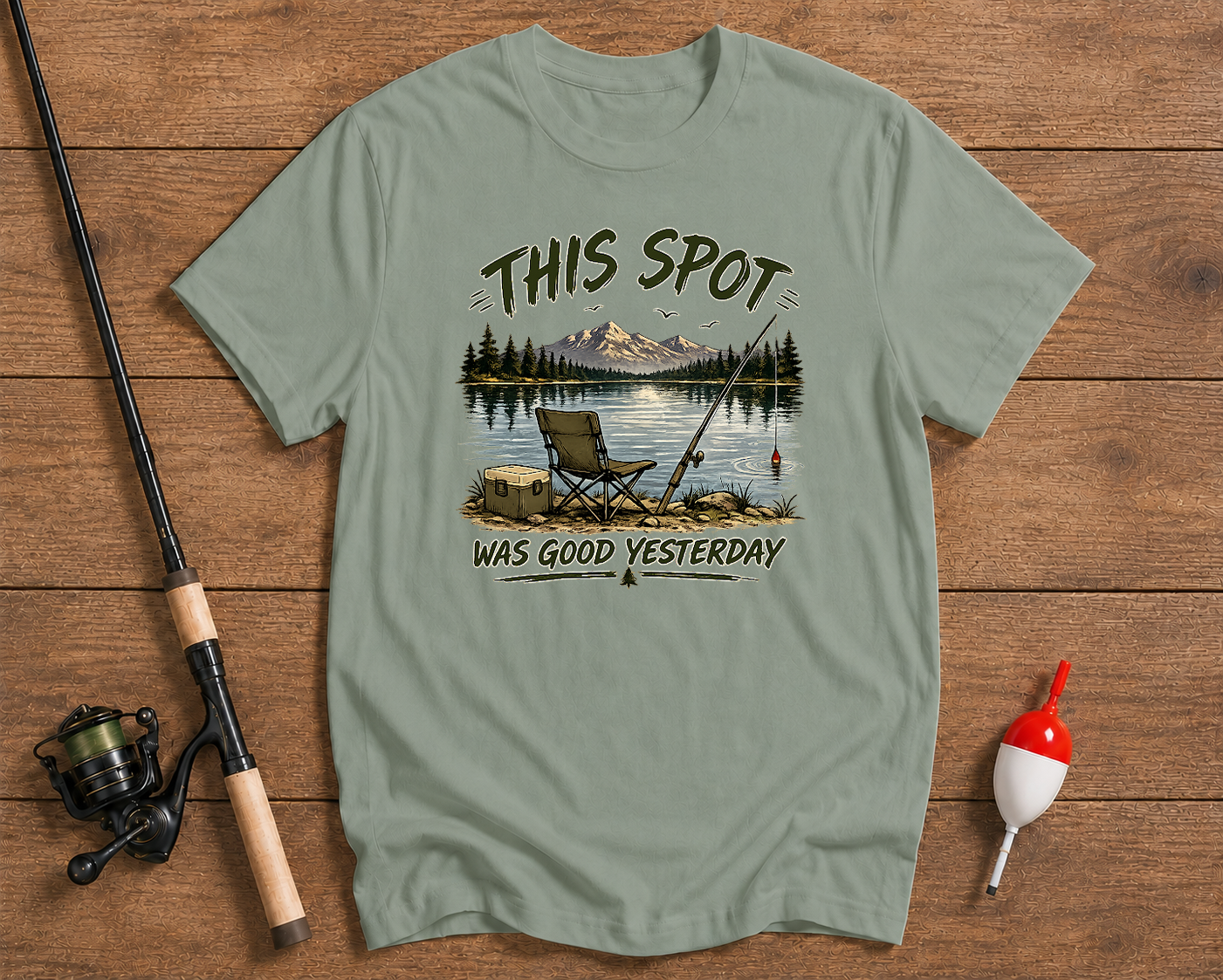 Sage green t-shirt with a fishing graphic and text on a wooden surface with fishing equipment.

