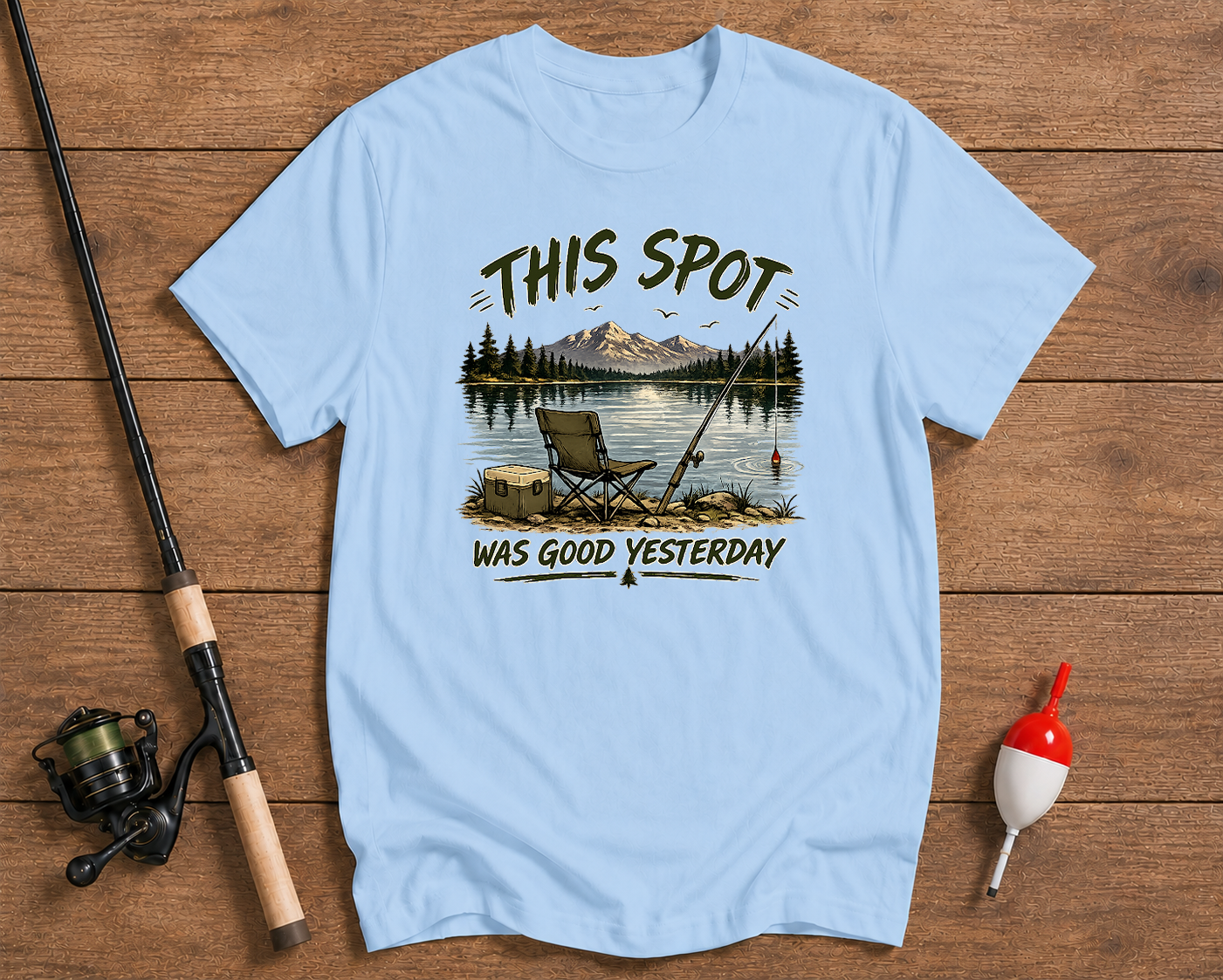 Light blue t-shirt with fishing graphic and text on a wooden surface with fishing equipment.

